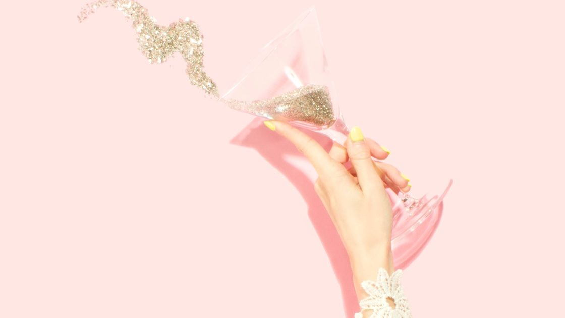 A pink background with a white woman's hand with painted yellow nails. The hand is holding a champagne glass full of gold glitter that is spilling out. There is a white lace bracelet around the wrist