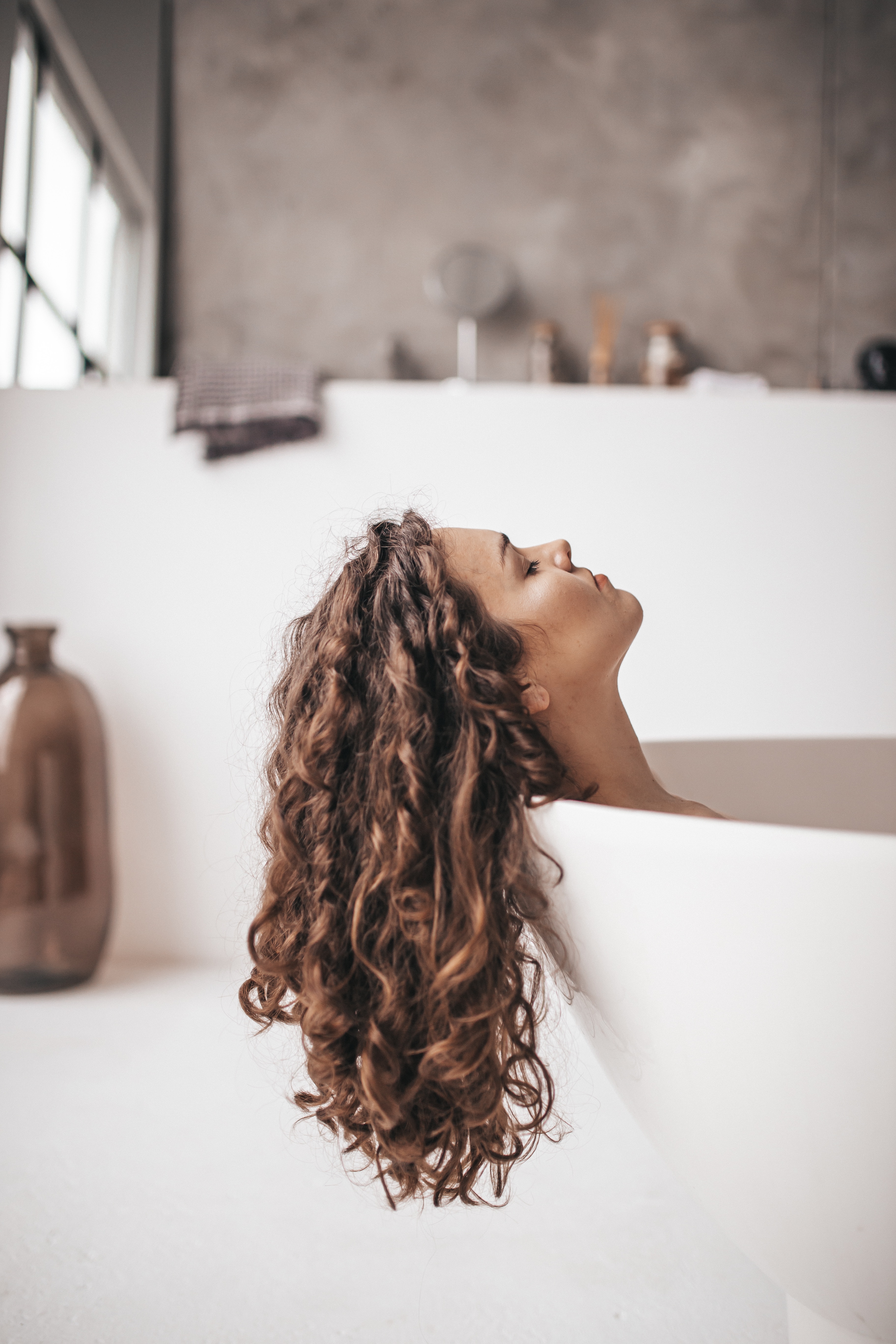 Woman reclines in a bathtub with long curly brown hair hanging down outside the tub