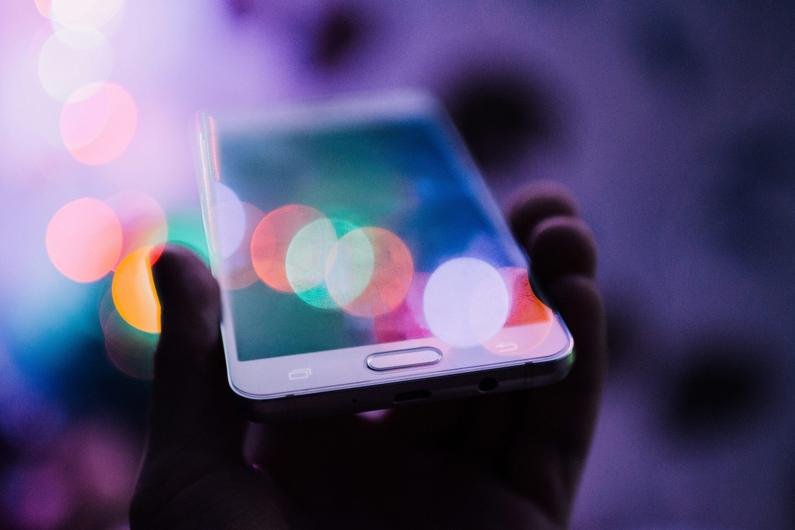 Dimly lit with purple hues. A hand holding a white smartphone up. Pastel colored ligt circles reflect on the phone screen.